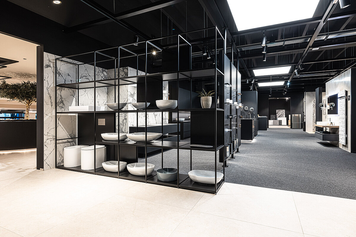 Ein moderner Ausstellungsraum zeigt Regale mit verschiedenen Sanitärkeramiken, darunter Waschbecken und Schalen in unterschiedlichen Formen und Farben. Der Raum ist stilvoll mit Marmorelementen an den Wänden und einem dunklen Teppichboden gestaltet und wird von natürlichem Licht erhellt.