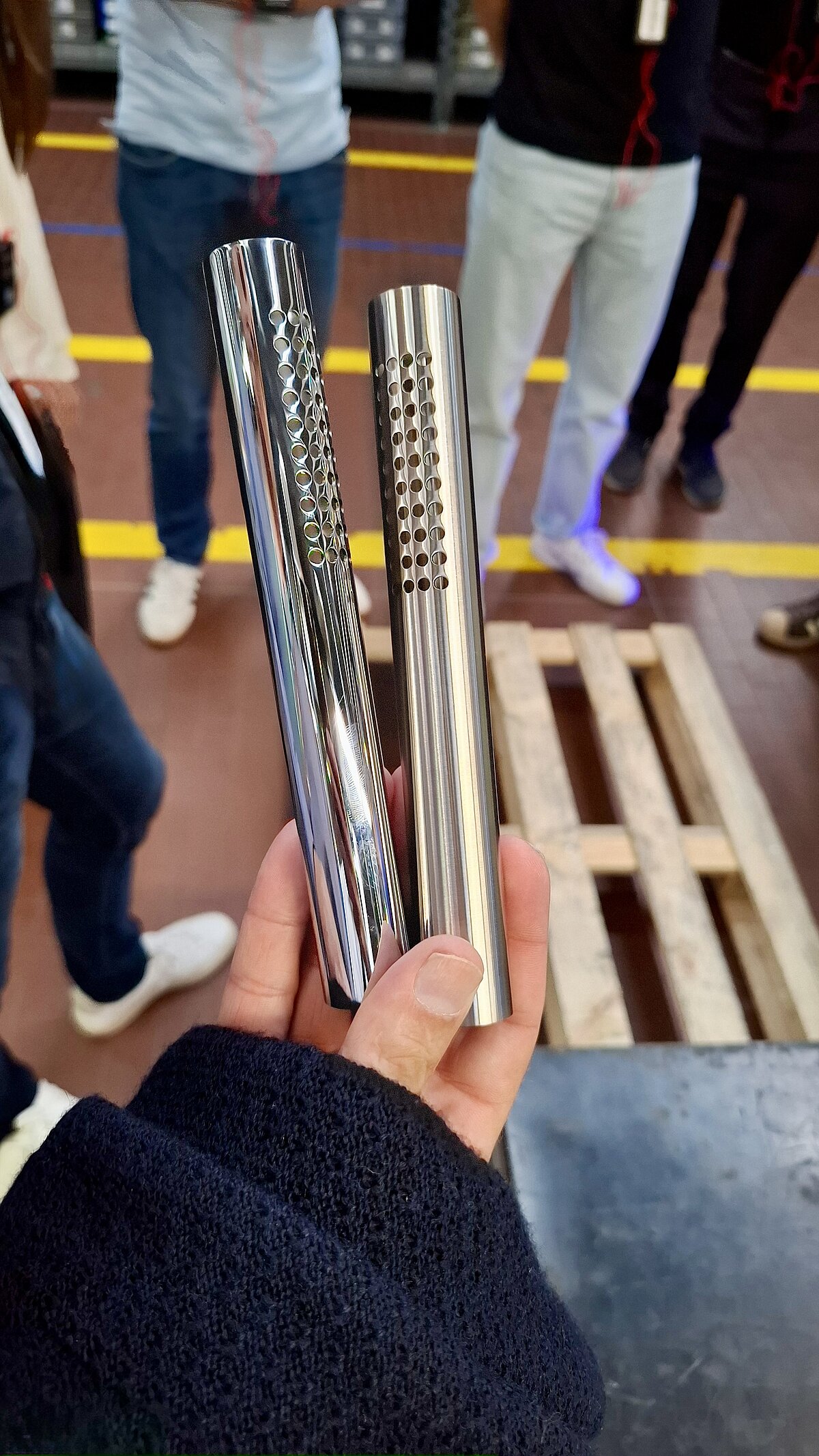 Eine Hand hält zwei glänzende, metallische Zylinder. Jeder Zylinder hat eine Reihe von Löchern entlang seiner Länge. Im Hintergrund stehen Menschen und ein Holzpalette liegt auf dem Boden.