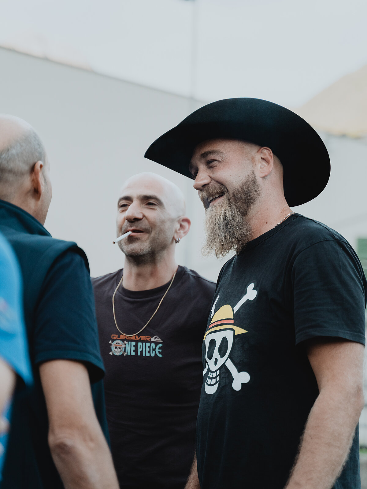 Drei Männer unterhalten sich im Freien. Einer trägt einen Cowboyhut und ein T-Shirt mit Totenkopf-Motiv. Ein anderer raucht eine Zigarette und trägt ein T-Shirt mit der Aufschrift "One Piece". Der dritte Mann ist nur von hinten zu sehen.
