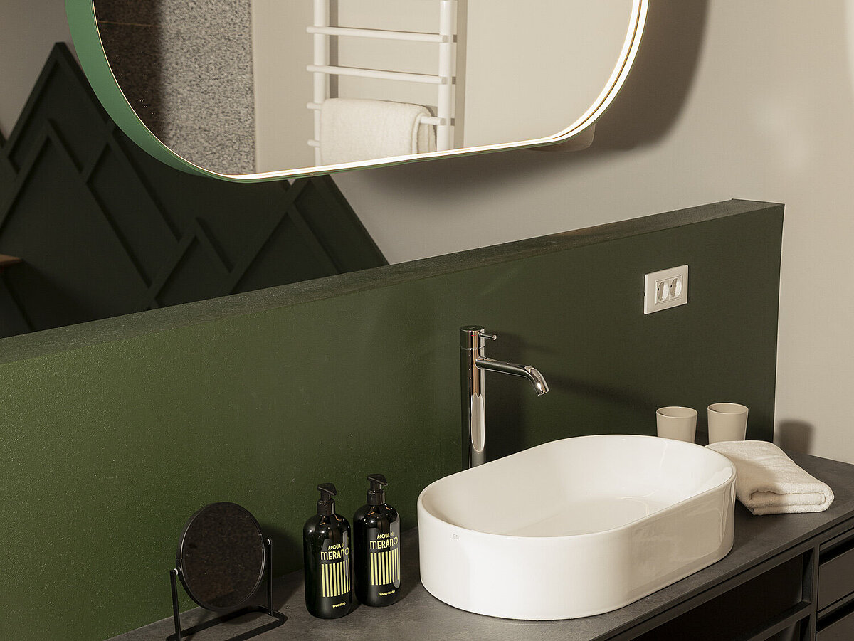 Ein moderner Waschraum mit einem ovalen Spiegel, der von weichem Licht umrahmt ist. Darunter befindet sich ein rechteckiges Waschbecken auf einer dunklen Arbeitsplatte. Neben dem Waschbecken stehen Pflegeprodukte und gefaltete Handtücher auf einem offenen, dunklen Regal.