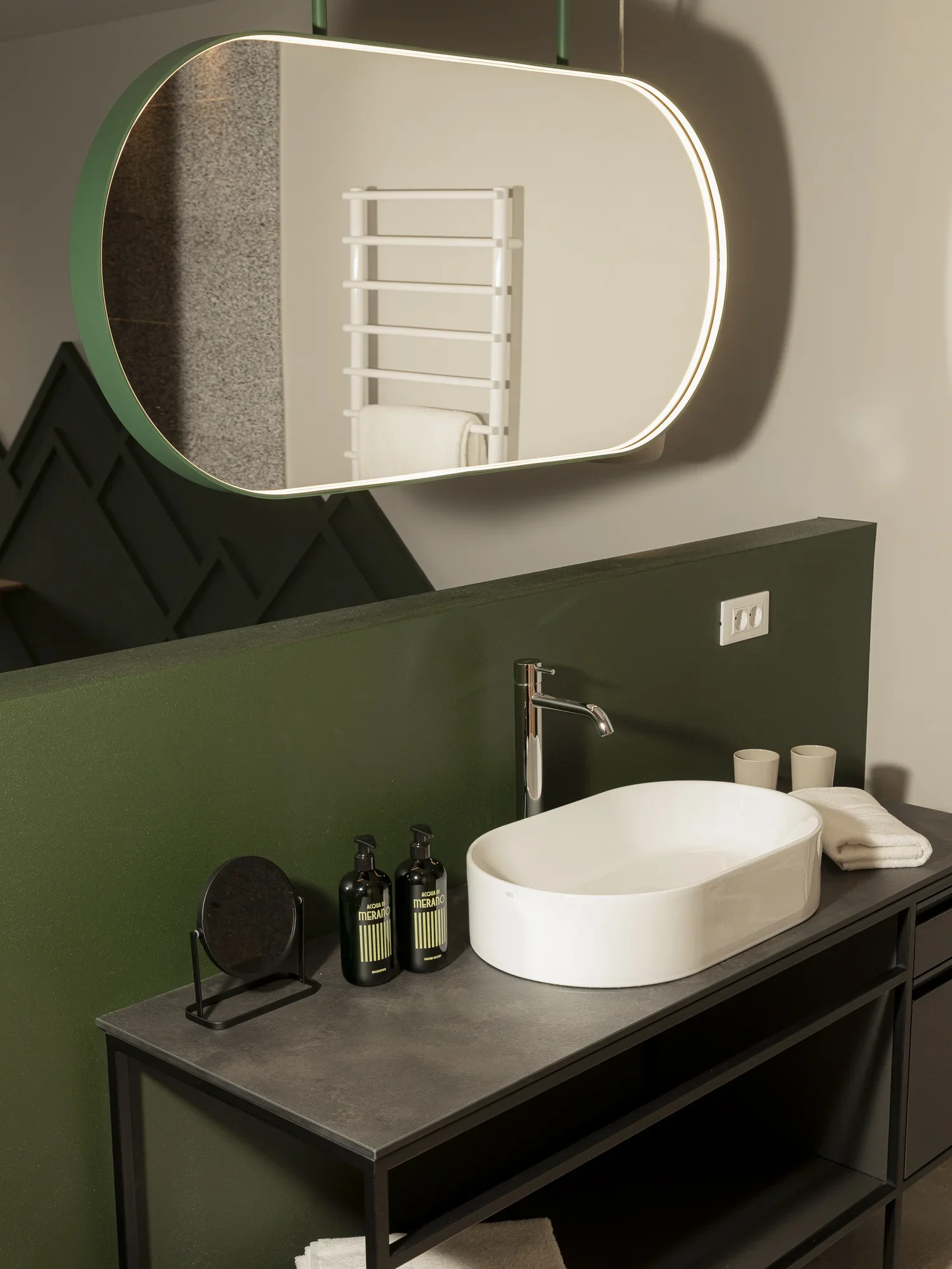 Ein moderner Waschraum mit einem ovalen Spiegel, der von weichem Licht umrahmt ist. Darunter befindet sich ein rechteckiges Waschbecken auf einer dunklen Arbeitsplatte. Neben dem Waschbecken stehen Pflegeprodukte und gefaltete Handtücher auf einem offenen, dunklen Regal.