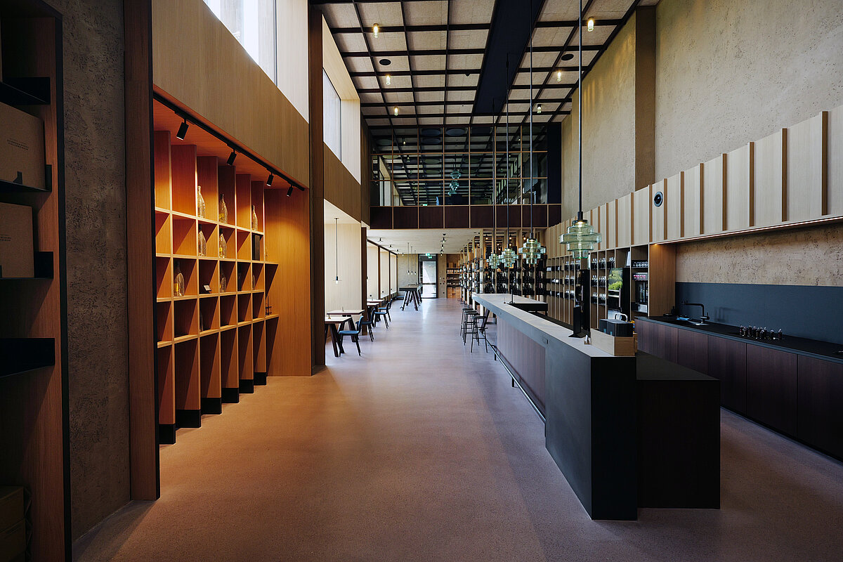 Moderner, minimalistischer Innenraum eines Cafés oder Restaurants mit hohen Decken. Links befinden sich hölzerne Regale, rechts eine lange Theke mit Hockern. Große Fenster lassen Tageslicht herein.