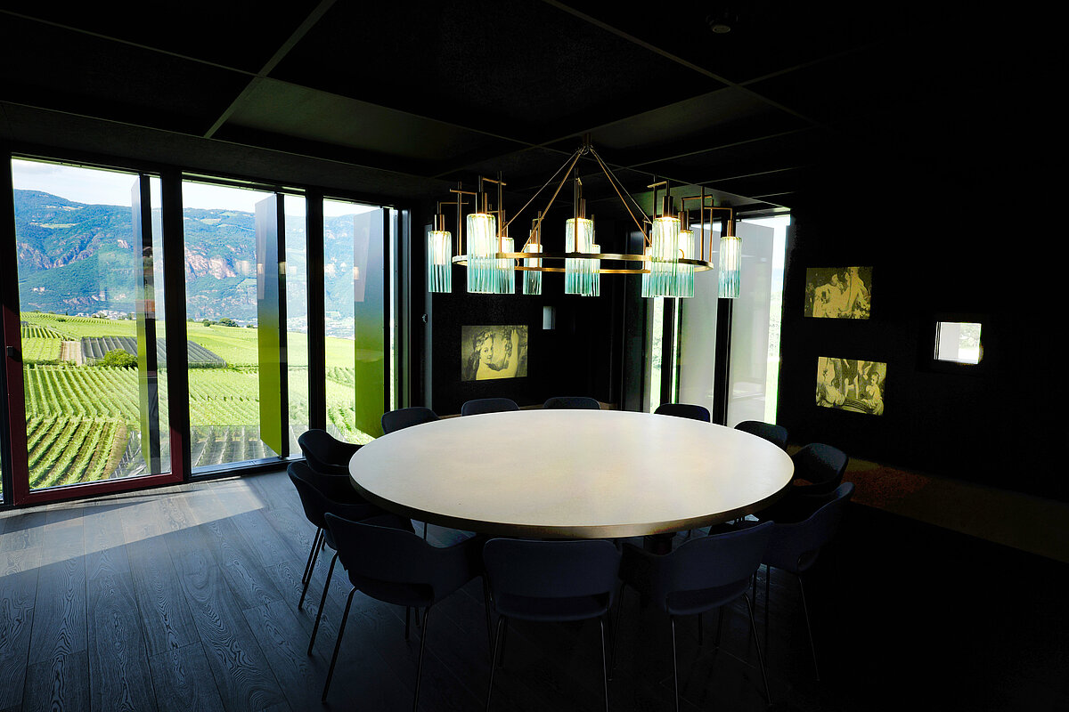 Runder Konferenztisch in einem modernen Raum, beleuchtet von einer Designer-Hängelampe. Durch große Fenster im Hintergrund ist eine Aussicht auf Weinberge und Hügel zu sehen. Wände sind dunkel mit beleuchteten Kunstwerken.