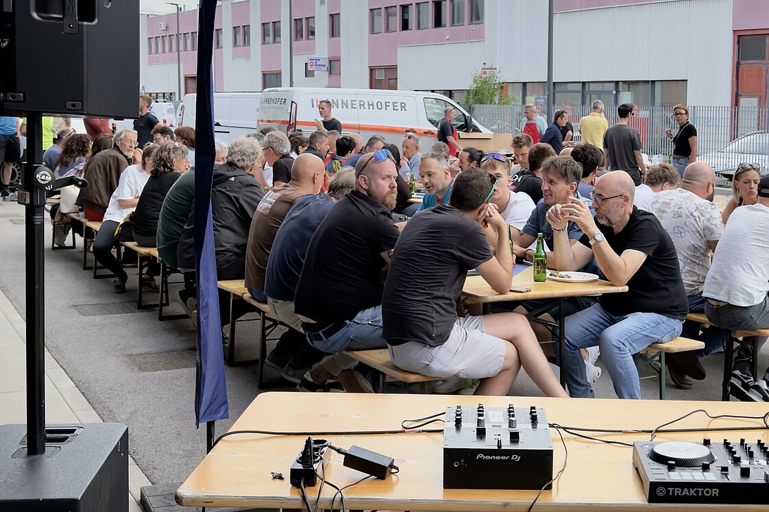 Menschen sitzen draußen an langen Holzbänken und essen. Im Vordergrund ist ein Tisch mit Audiogeräten, im Hintergrund ein großes modernes Gebäude. Der Himmel ist bewölkt.