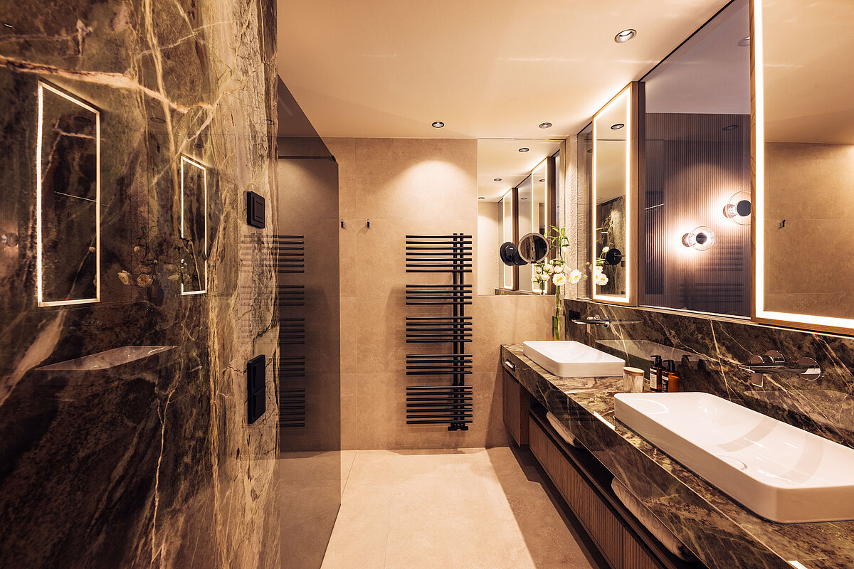 Modernes Badezimmer mit großen Spiegeln und zwei weißen Waschbecken auf einer dunklen Marmorplatte. Die Wände sind in warmen Beigetönen gehalten. Eine Glasduschkabine mit Regenduschkopf und stilvollen schwarzen Armaturen ergänzt den eleganten Raum. Beleuchtungskörper verstärken die luxuriöse Atmosphäre.