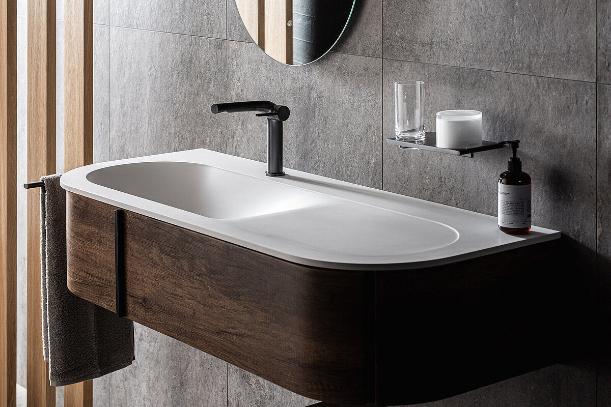 Modernes Badezimmer mit ovalem Spiegel, weißem Waschbecken und schwarzem Wasserhahn. Dunkles Holz und graue Kacheln dominieren das Design. Neben dem Waschbecken stehen ein Seifenspender und ein kleines Glas. Ein Handtuch hängt an der Seite des Waschbeckens.