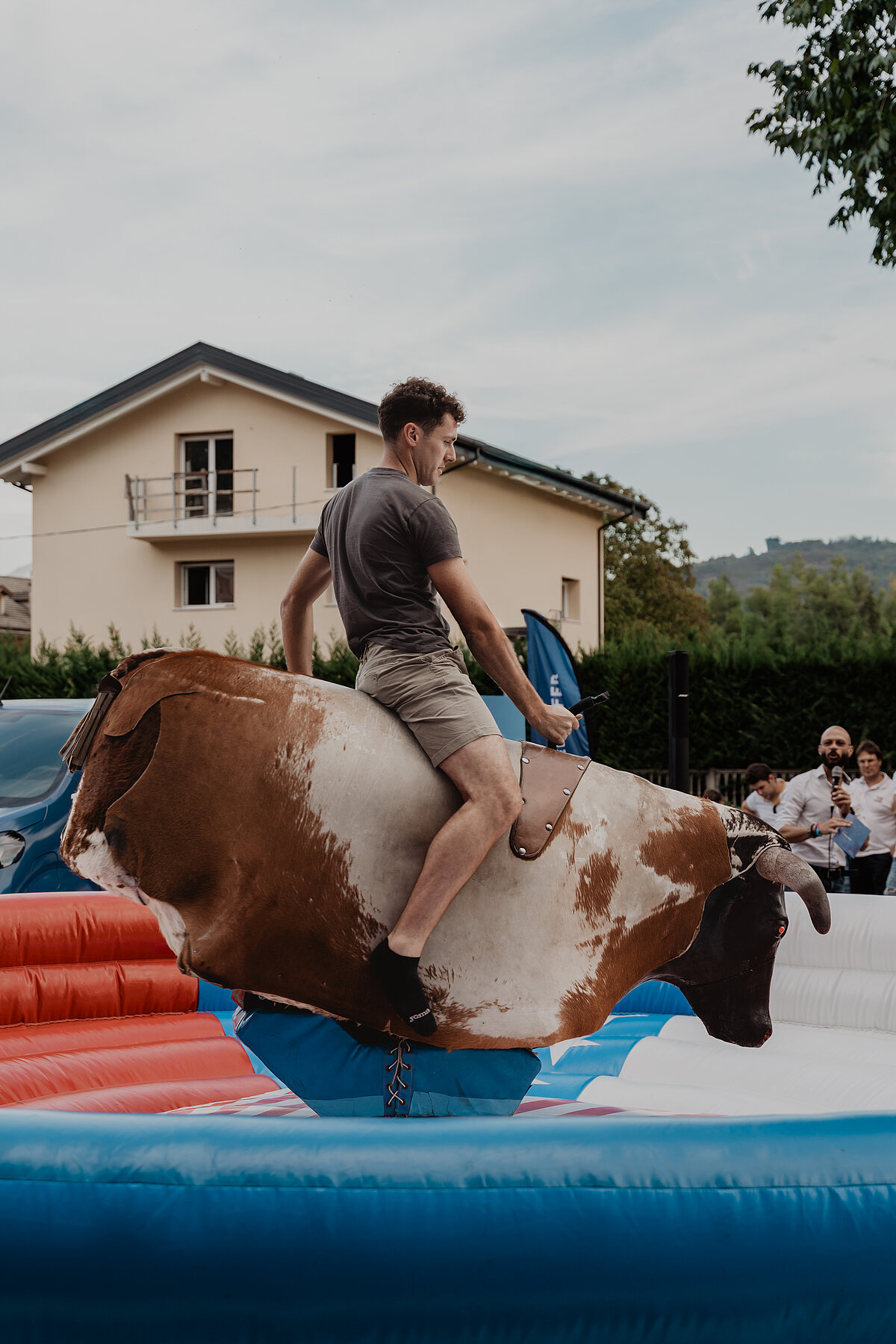 Ein Mann reitet auf einem mechanischen Bullen in einem aufblasbaren Bereich. Im Hintergrund ist ein Haus und eine kleine Menschenmenge zu sehen, die zuschaut. Der Himmel ist bewölkt.