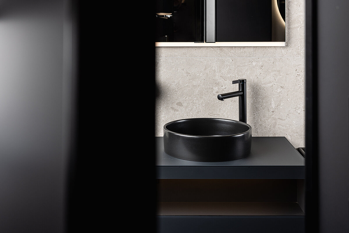 Ein modernes Badezimmer mit einem schwarzen, runden Waschbecken und einem minimalistischen Wasserhahn auf einer grauen Arbeitsplatte. Im Hintergrund ist eine helle, geflieste Wand zu sehen.
