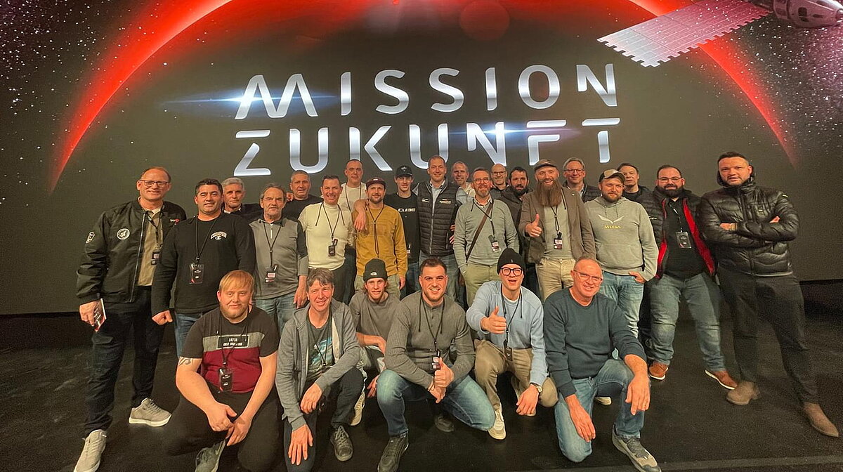 Eine Gruppe von Menschen steht vor einem großen Bildschirm mit der Aufschrift „Mission Zukunft“, das einen futuristischen Hintergrund zeigt. Alle blicken in die Kamera und lächeln.