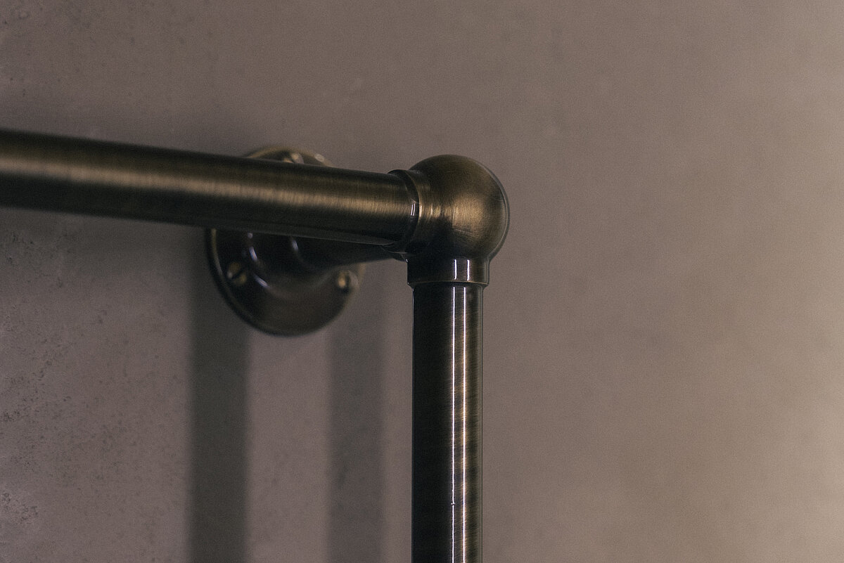 Nahaufnahme einer Metallrohrverbindung in einem rechten Winkel, montiert an einer grauen Wand. Die Oberfläche des Rohrs hat eine dunkelbronzene Farbe und wirkt matt.
