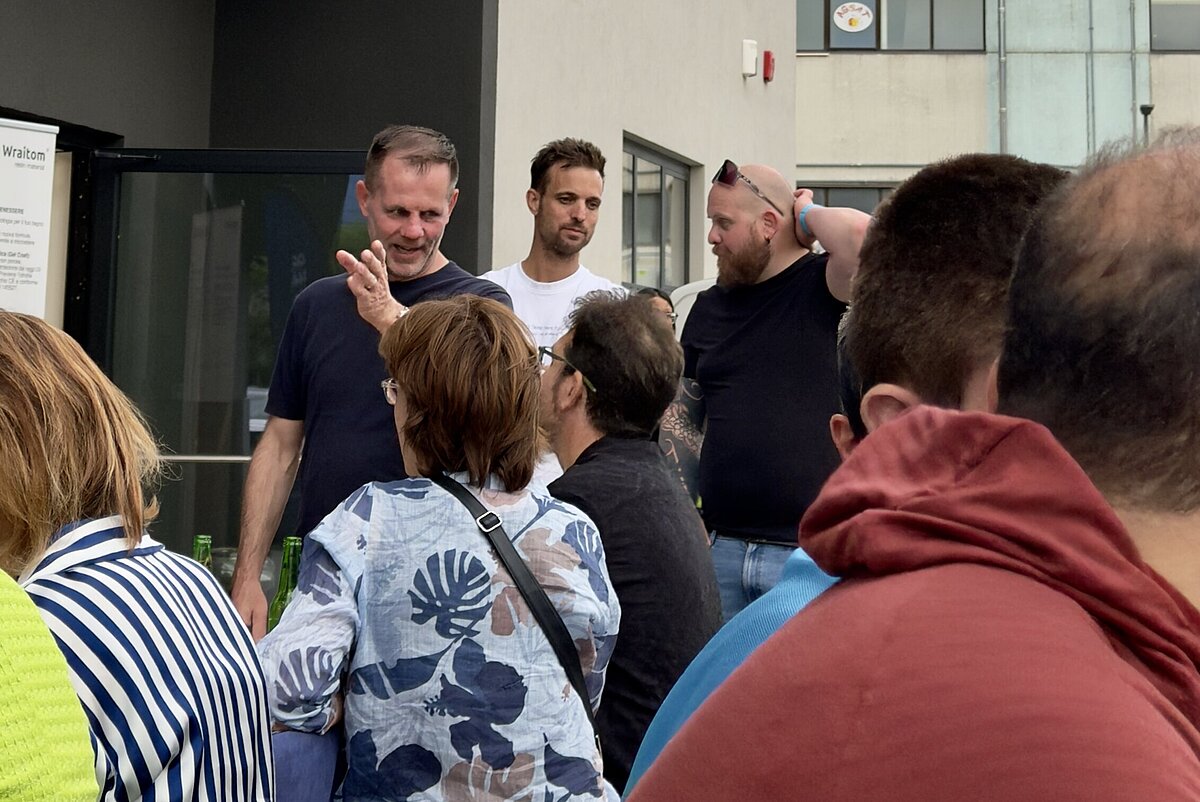 Eine Gruppe von Menschen steht vor einem modernen Gebäude mit der Aufschrift "Linsenhöfer". Sie unterhalten sich, während sie in einer Schlange zu stehen scheinen. Der Himmel im Hintergrund ist leicht bewölkt.