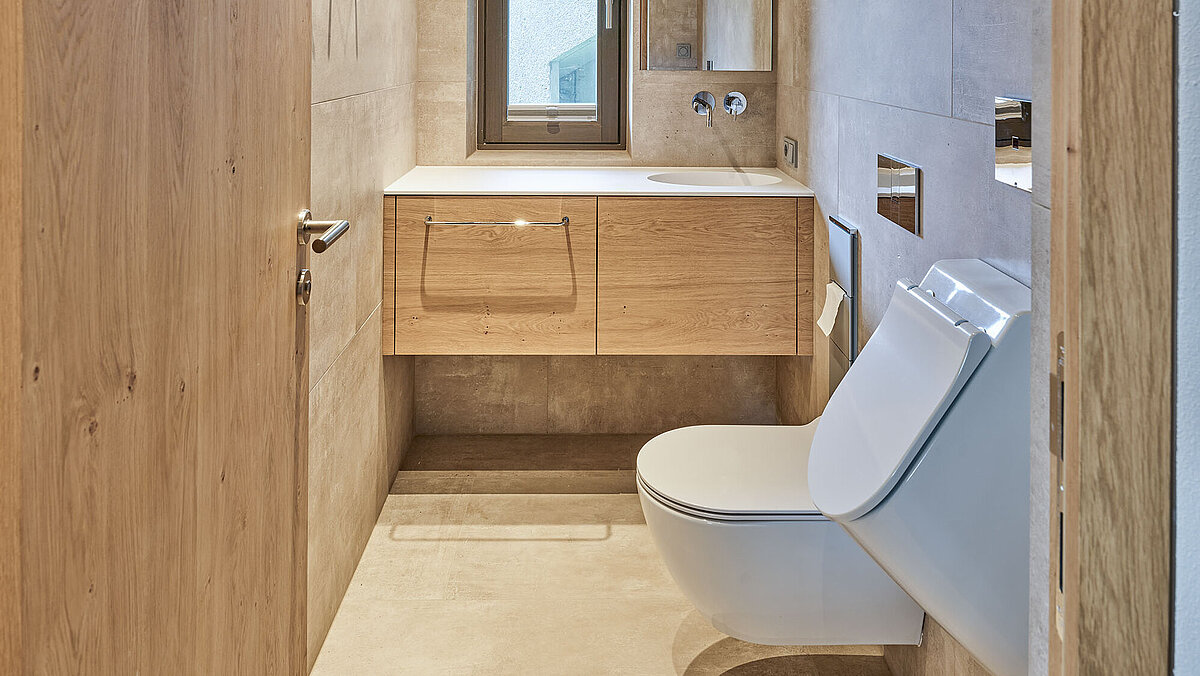Modernes Badezimmer mit Holzverkleidung. Auf der rechten Seite befindet sich eine Wandtoilette, links ein Holzwaschbecken mit Spiegel darüber. Durch ein schmales Fenster fällt Tageslicht ein. Deckenstrahler beleuchten den Raum.