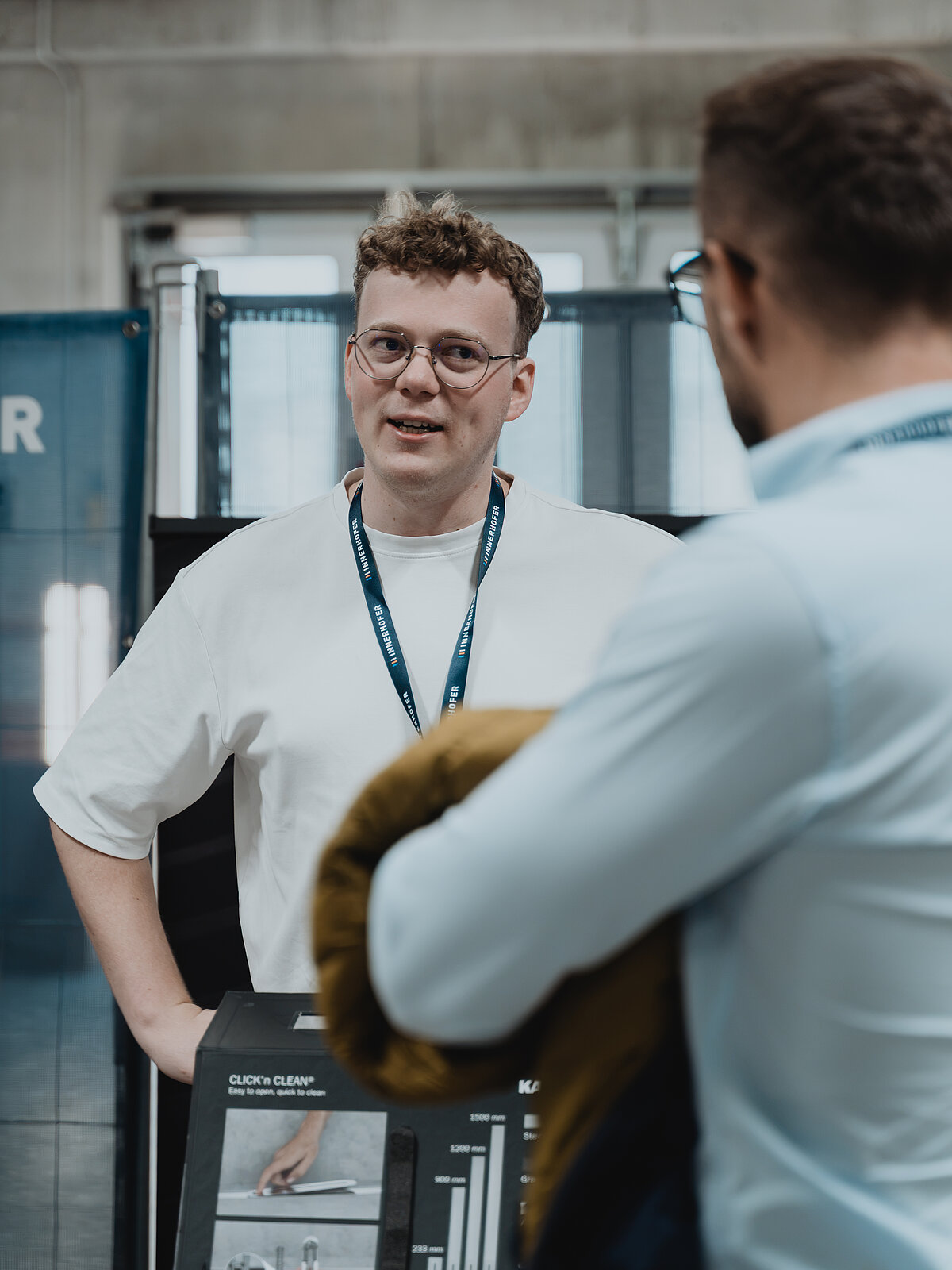 Eine Person mit Brille und Namensschild spricht mit einer anderen Person, die den Rücken zur Kamera hat. Sie befinden sich in einem modernen, industriellen Raum. Es sind Informationsposter oder Schilder im Hintergrund zu sehen.