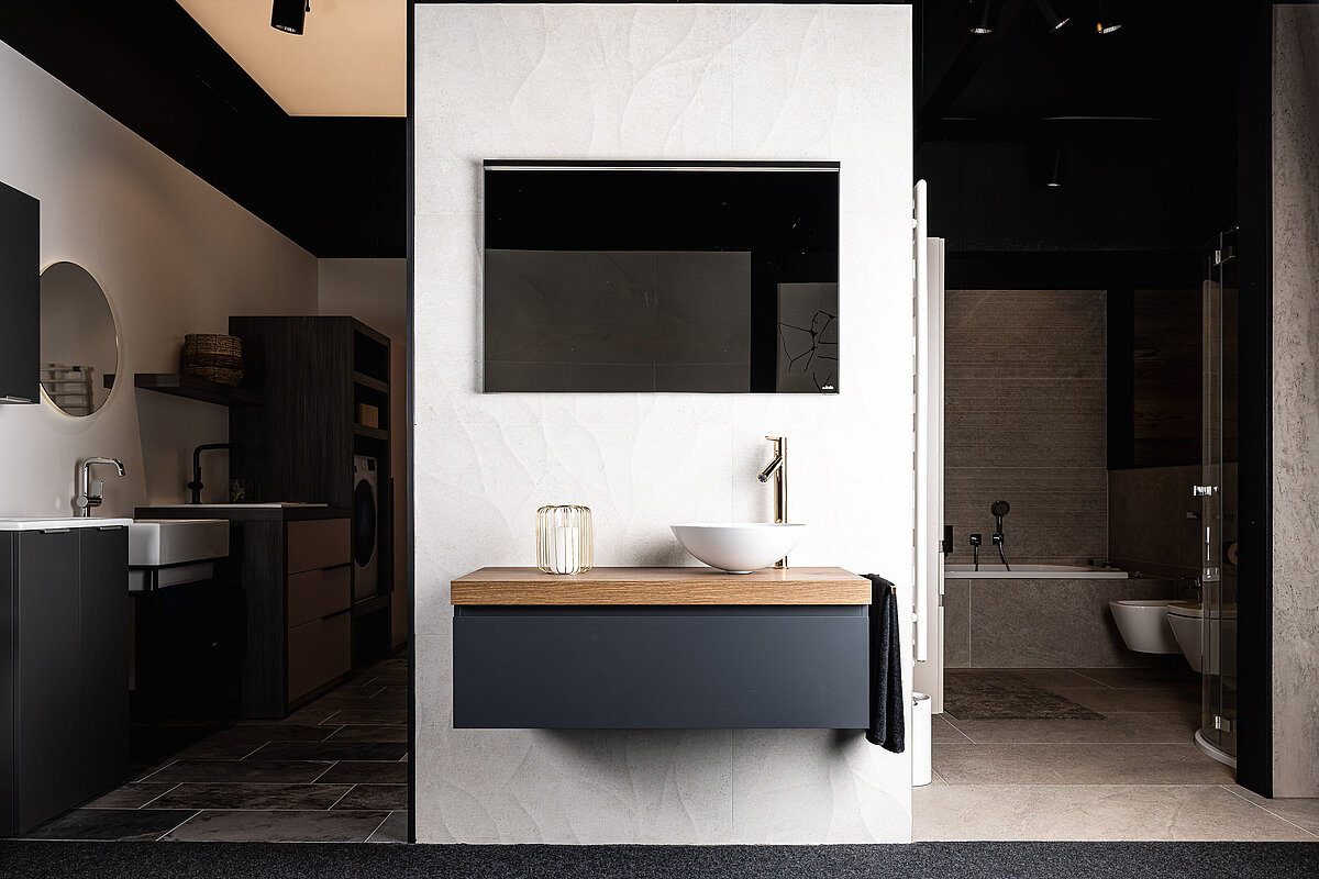 Ein modernes Badezimmer mit einem großen Spiegel, einem Waschbecken auf Holzplatte und schwarzen Schränken. Dahinter ist ein offenes weiteres Bad mit freistehender Badewanne und dunklen Fliesen sichtbar. Elegante, minimalistische Gestaltung mit hellen und dunklen Farben.