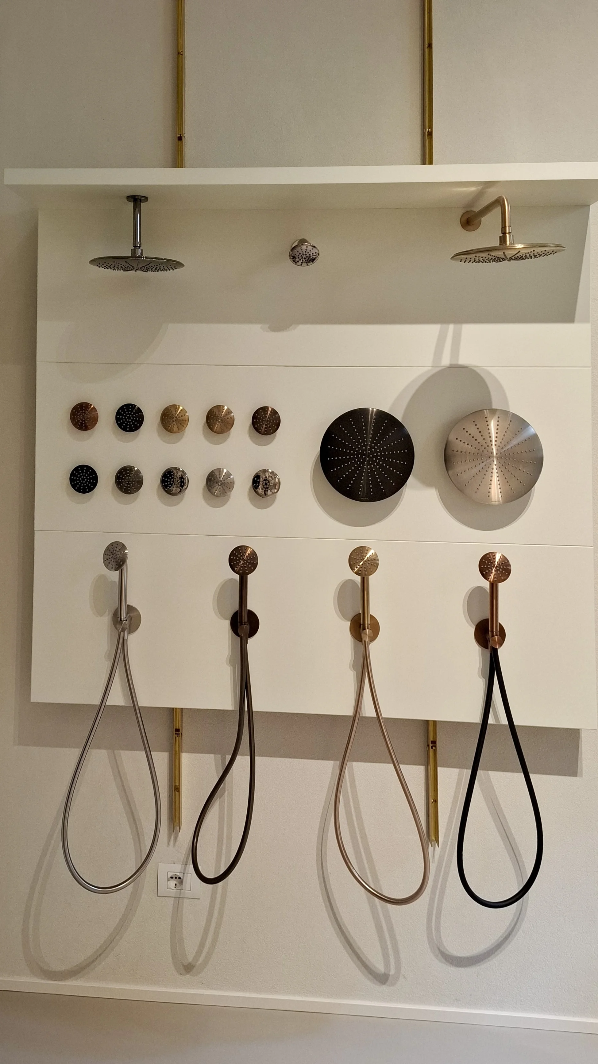 Duscharmaturenausstellung mit verschiedenen Brauseköpfen und Bedienelementen in unterschiedlichen Oberflächen und Farben, die an einer weißen Wand montiert sind. Darunter sind flexible Duschschläuche in verschiedenen Farbtönen angeordnet.