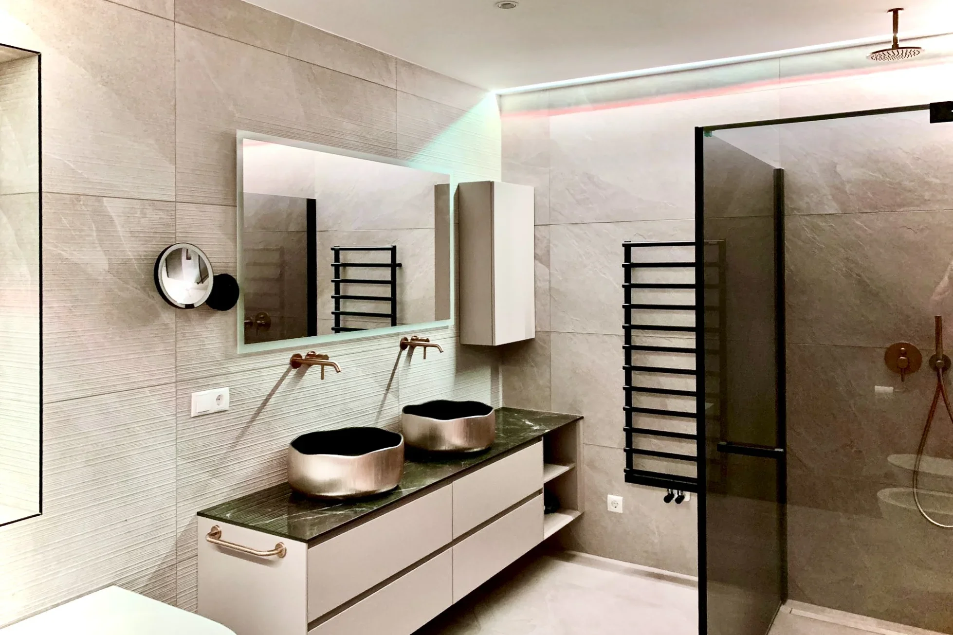 Modernes Badezimmer mit zwei Waschbecken auf einem langen Waschtisch, einem großen Spiegel darüber und einer begehbaren Dusche mit Glaswand. Die Wände und der Boden sind mit hellen Fliesen verkleidet. Ein Handtuchwärmer ist an der Wand montiert.