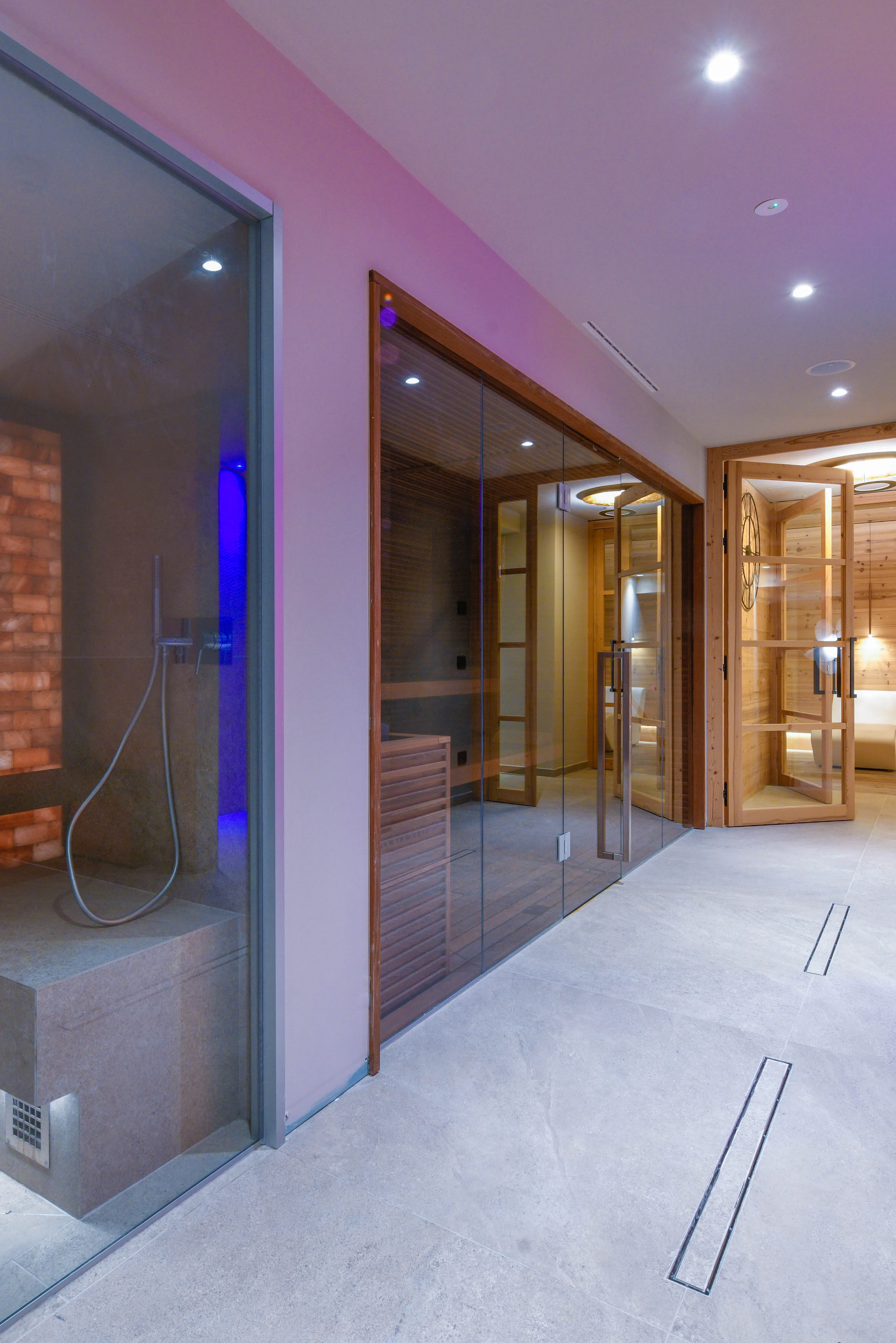 Ein moderner Spa-Bereich mit mehreren Saunen. Links ist ein Dampfbad mit Ziegelwand und Dusche sichtbar. Rechts sind ein Infrarotraum und eine Sauna mit Holzbänken zu sehen. Der Boden ist aus hellem Stein. Es gibt sanfte Deckenbeleuchtung.