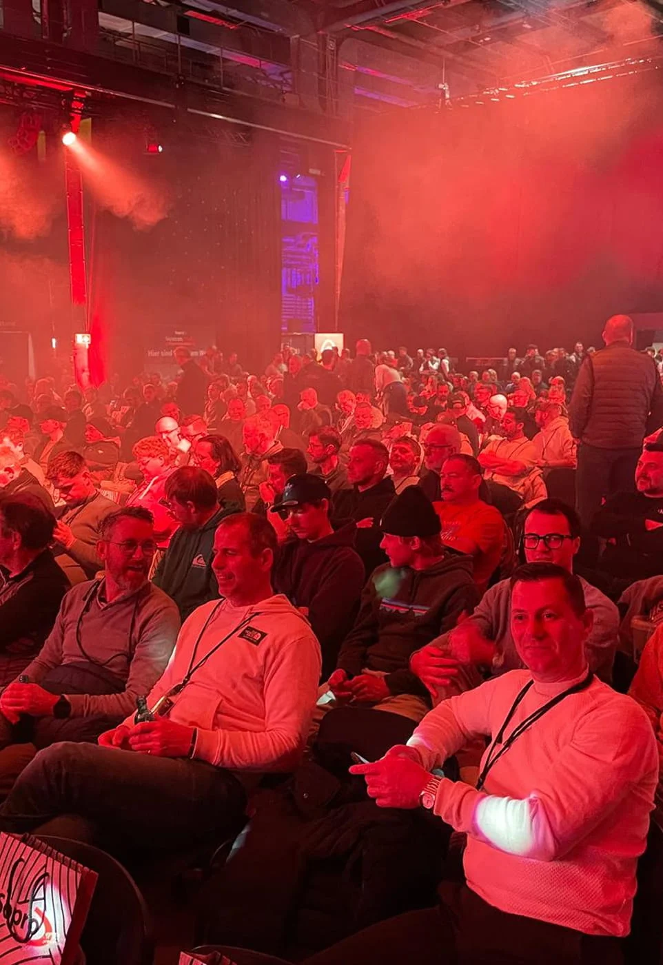 Eine große Anzahl von Menschen sitzt in einem Veranstaltungsraum unter roten Lichtern. Die Personen scheinen auf eine Präsentation oder Rede zu warten. Die Stimmung wirkt gespannt und erwartungsvoll.