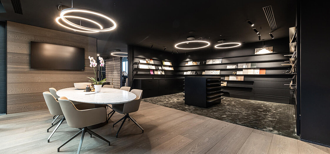 Modernes Büro mit schwarzem Interieur, runden Deckenleuchten und einem großen, weißen Konferenztisch mit sechs Stühlen. Im Hintergrund befinden sich Regale mit Büchern und Zeitschriften. Ein Blumenarrangement steht auf dem Tisch.